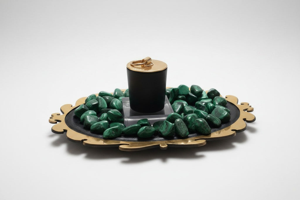 Citrine Home Malachite candle in a black vessel with a gold lid, surrounded by green malachite stones on a tray.