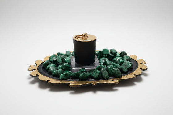 Citrine Home Malachite candle in a black vessel with a gold lid, surrounded by green malachite stones on a tray.