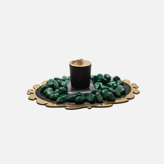 Citrine Home Malachite candle in a black vessel with a gold lid, surrounded by green malachite stones on a tray.
