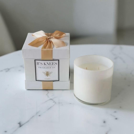 Citrine Home Veneto white scented candle in a glass jar next to a gift box with a gold ribbon on a marble table.